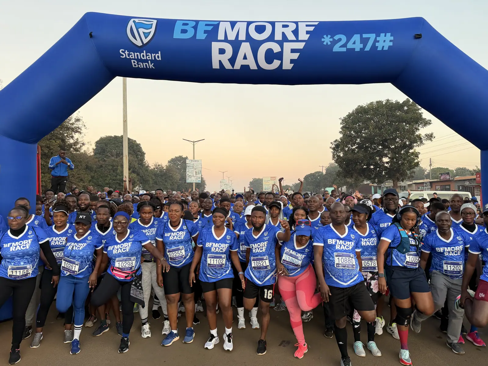 Runners in blue shirts crowd the start under the Be More Race arch